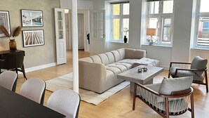 Amplia sala de estar con un sofá seccional color beige, una mesa de centro y un comedor con mesa oscura y sillas claras. Hay ventanas grandes con cortinas, una puerta blanca y cuadros en las paredes.