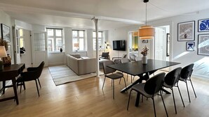 Una sala de estar moderna con sofá, mesa de comedor y sillas. Cuenta con ventanas amplias y un televisor montado en la pared.