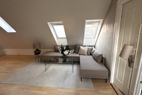 A modern loft apartment with a glass coffee table, a grey sofa, and a large window providing natural light.