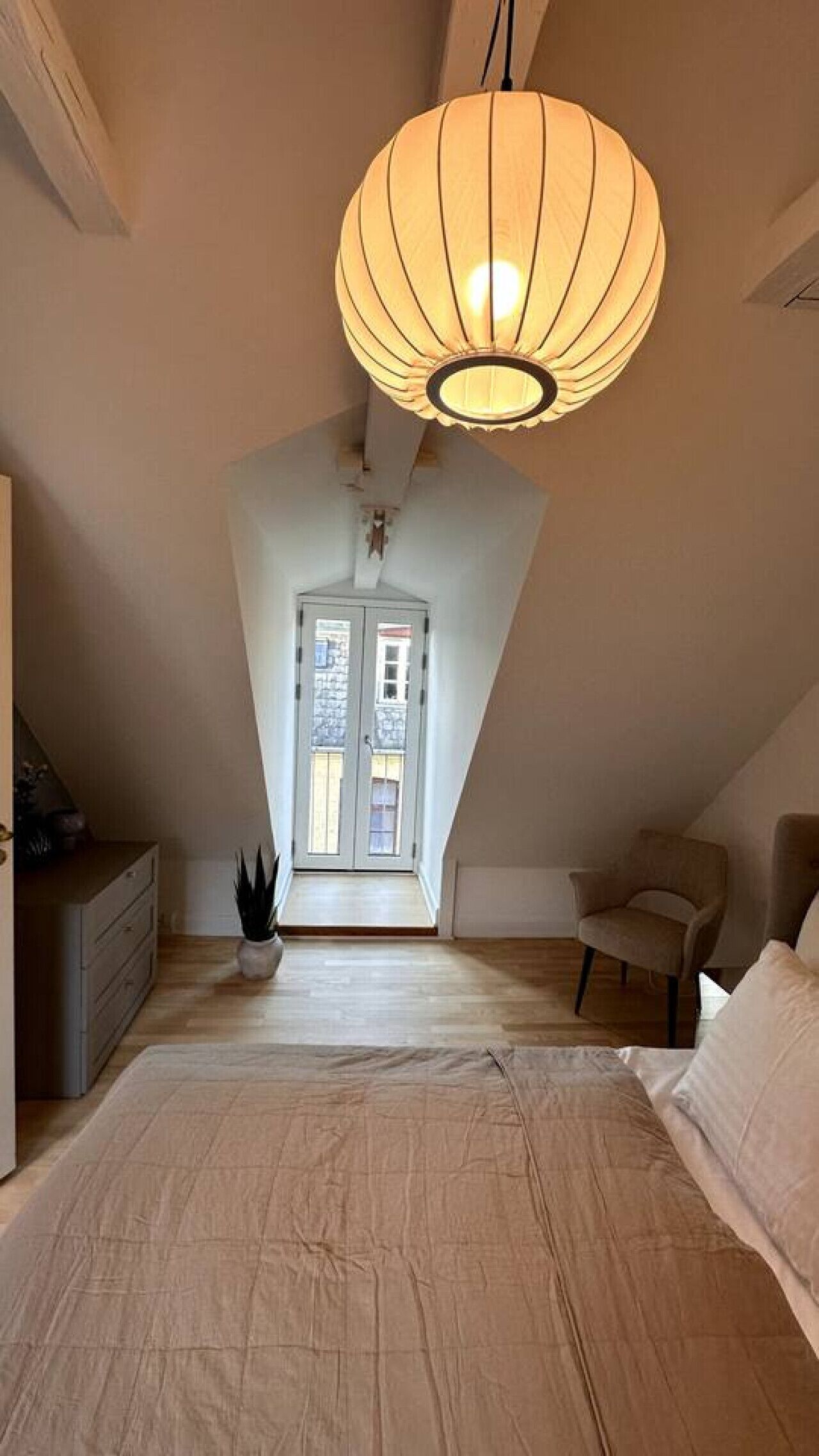 Una camera da letto con soffitto spiovente, un letto grande, una lampada a sospensione, una sedia e vista sulla strada attraverso una porta.