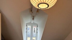 A bedroom with a sloped ceiling, a large bed, a hanging light fixture, a chair, and a view of the street through a door. - Løvstræde baghus 4 th top floor (København)
