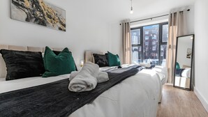 A modern hotel room with a large bed, multiple pillows, a black throw blanket, and a view of the city through large windows.