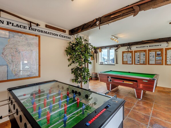 Une table de billard, une table de baby-foot, une carte et une plante dans une pièce aux poutres apparentes et au sol carrelé.