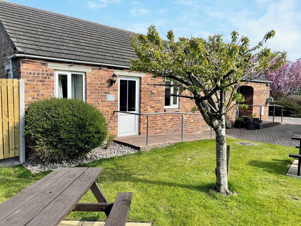 A brick building with a tiled roof, a wooden fence, a picnic table, and a tree.