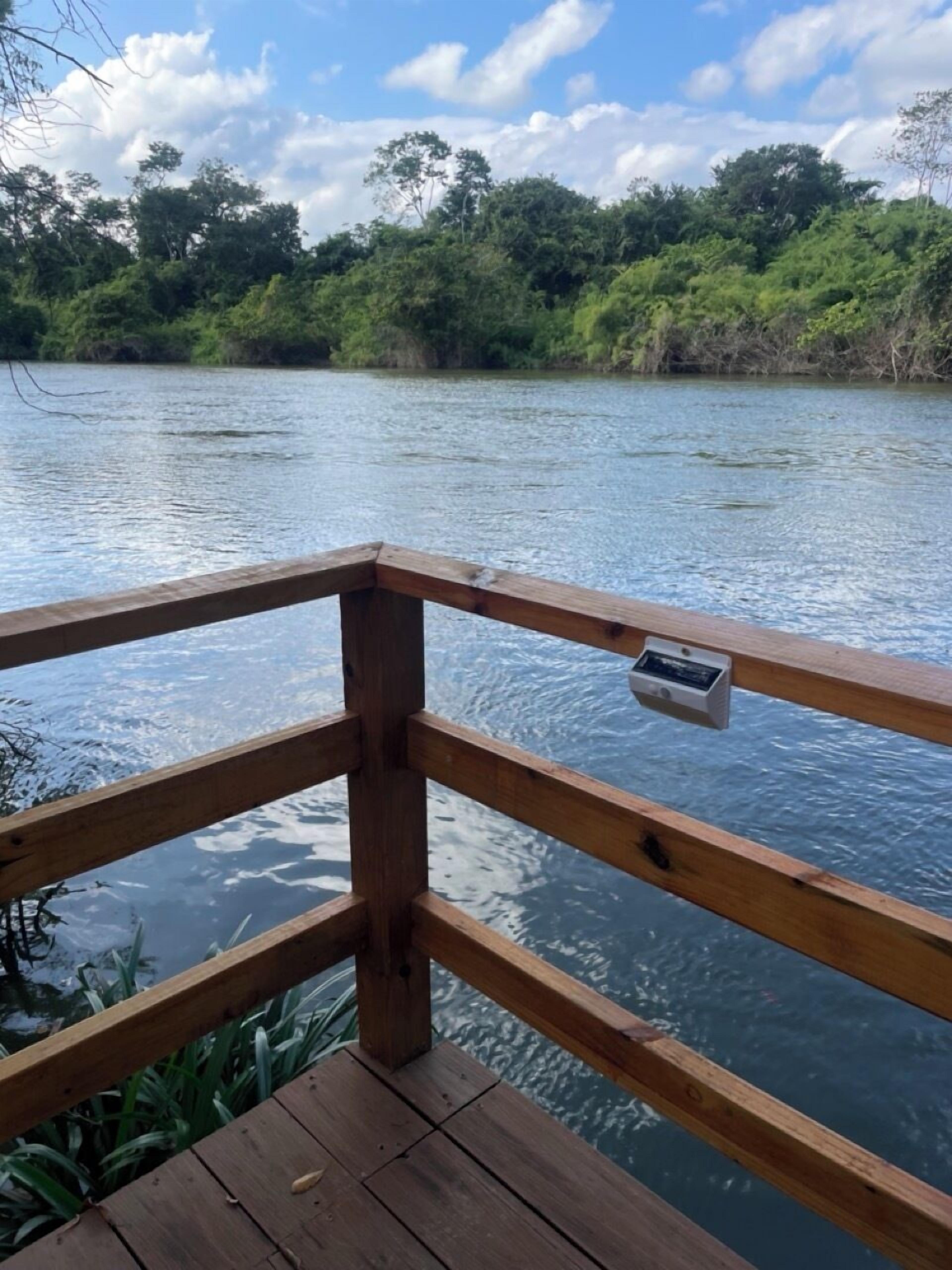 Une terrasse en bois dotée d’une balustrade, surplombant une étendue d’eau calme, et entourée d’une végétation luxuriante et d’arbres.