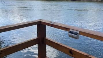 Une terrasse en bois dotée d’une balustrade, surplombant une étendue d’eau calme, et entourée d’une végétation luxuriante et d’arbres.