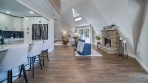 Una cocina moderna con armarios blancos y un refrigerador de acero inoxidable. Hay una zona de comedor con un taburete de bar y un sofá azul. Se ve una chimenea de piedra con un cartel que dice 