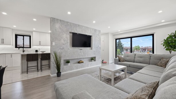 A modern living room with a grey sofa, a flat-screen TV mounted on a marble wall, and a kitchen area with white cabinets and a bar counter. - Luxury 3 BDRM Home In Toronto Ontario (Toronto)