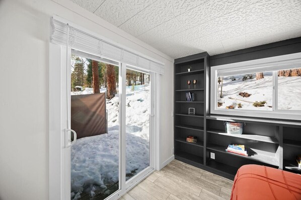 A room with a sliding glass door leading to a snowy outdoor area, a bookshelf with various items, and a window with a view of the outside.