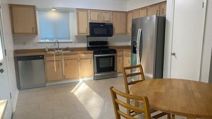 Una cocina con armarios de madera, electrodomésticos de acero inoxidable, lavavajillas, microondas, horno y refrigerador. Hay una mesa de comedor de madera con sillas.
