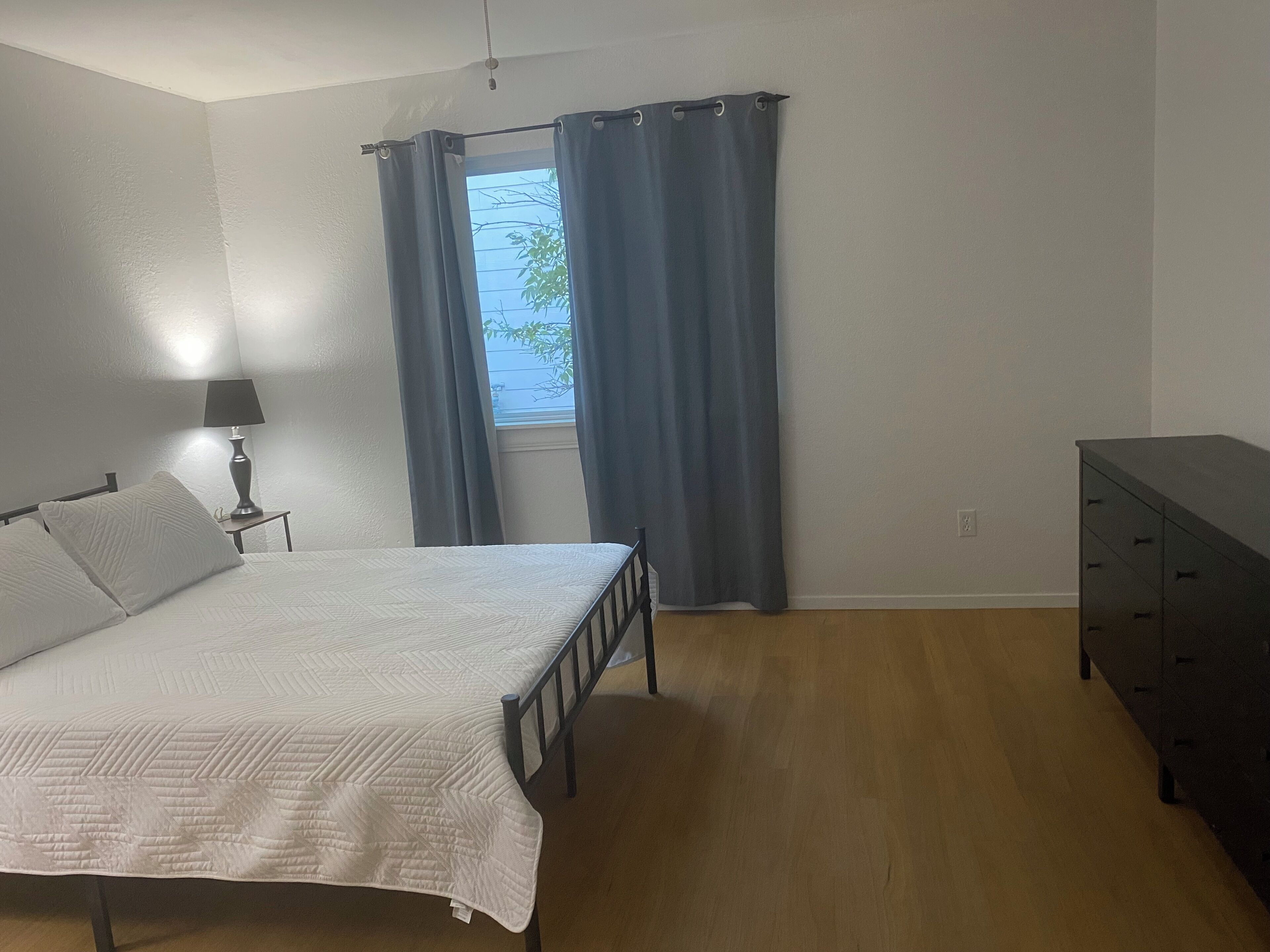 Une chambre à coucher avec un lit, une table de chevet avec une lampe, une commode et une fenêtre avec des rideaux.