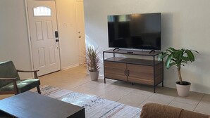 Una sala de estar con ventilador de techo, televisor sobre un soporte de madera, sofá, mesa de centro y dos plantas en macetas.