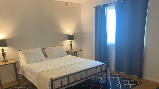 Un dormitorio con cama, dos mesitas de noche con lámparas, una ventana con cortinas y una alfombra estampada.