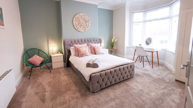 A bedroom with a tufted headboard bed, a green chair, a desk, and a large window. - Shoreline Angel Apartments (South Shields)