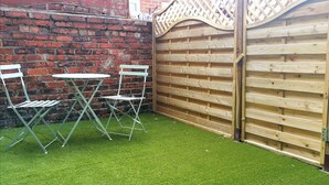 A small outdoor area with artificial grass, two white metal chairs, and a wooden fence. - Shoreline Angel Apartments (South Shields)