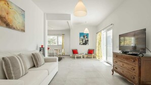 Una sala de estar moderna con un sofá blanco, un aparador de madera y un televisor. Se observa un comedor con sillas y una mesa al fondo.
