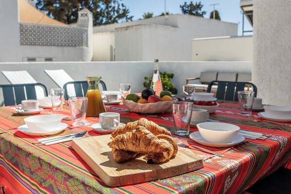 A breakfast setup with croissants on a wooden board, a fruit bowl, and various breakfast items on a colorful tablecloth.