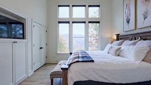 A bedroom with a large window, a bed with a quilt, a bench, and a TV mounted on a cabinet.