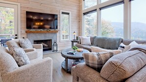 A cozy living room with a fireplace, comfortable seating, and large windows offering a view of the outdoors.