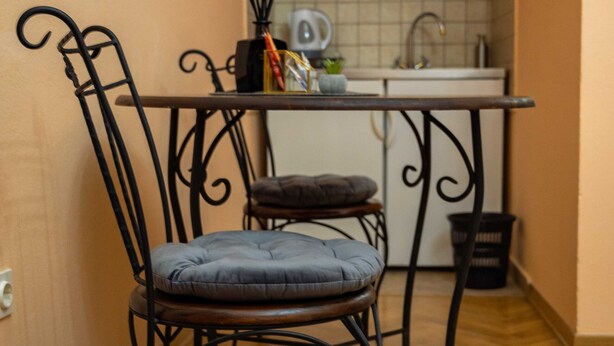 Un pasillo estrecho con suelo de madera, una silla metálica con cojín, una pequeña mesa con candelabro y una zona de cocina con armarios y un lavabo.