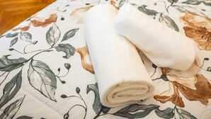 Una cama bien tendida con una colcha estampada y dos toallas enrolladas, una puerta de madera con persianas, y una mesita con una lámpara.