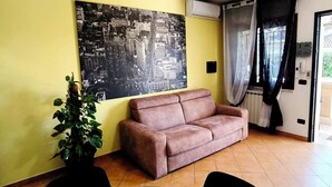 Una sala de estar con un sofá, una mesa de comedor, un ventilador de techo, un cuadro grande de un paisaje urbano y una planta en maceta.