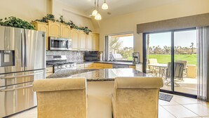 A modern kitchen with stainless steel appliances, a large island with bar stools, and a view of a golf course through the sliding glass doors. - Community Pools & Golf Views: Palm Desert Condo! (Palm Desert)