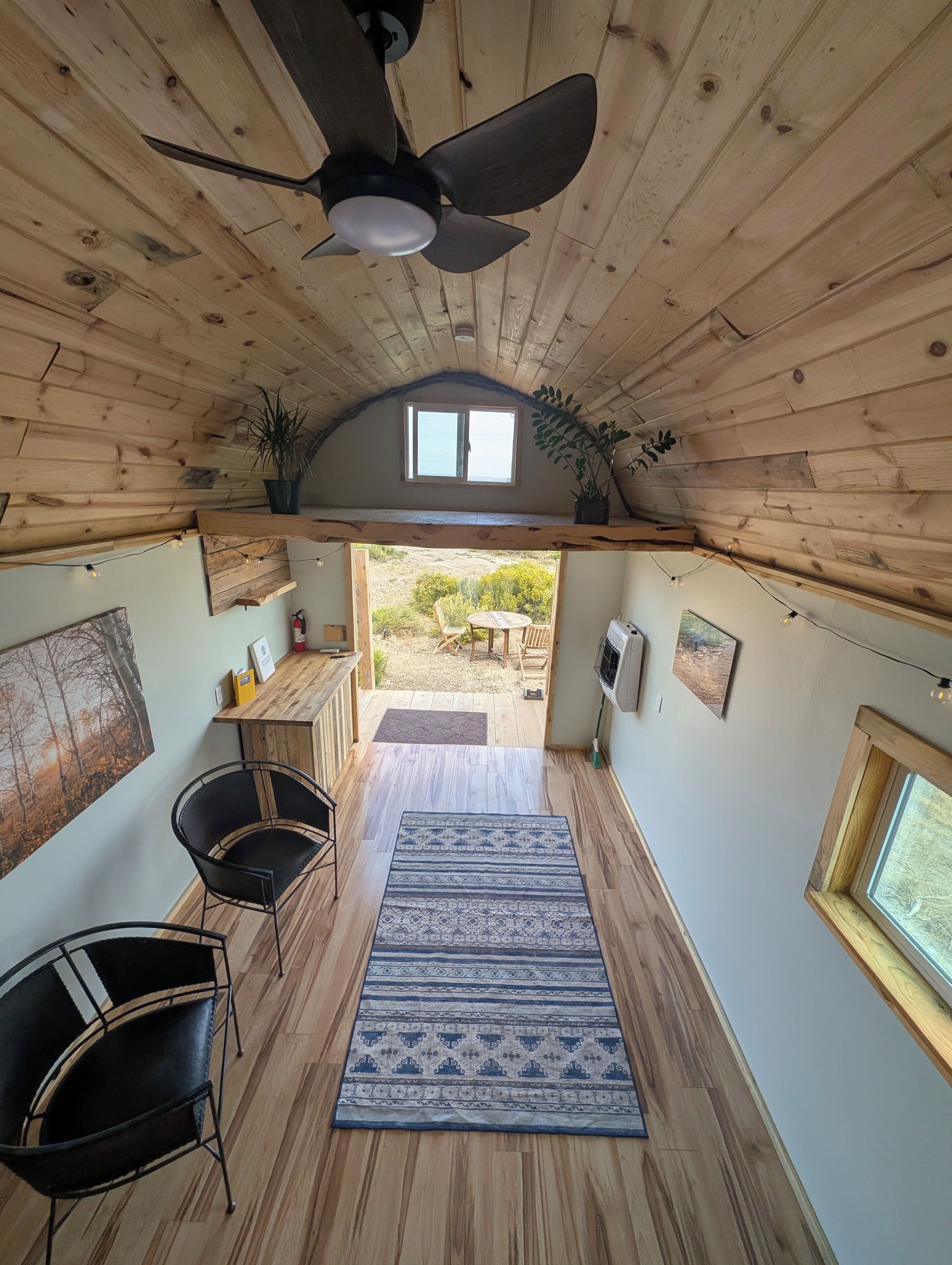 Une pièce chaleureuse avec des murs et un plafond en bois, un ventilateur de plafond, un coin repas avec des chaises et une table, et une vue sur l’extérieur.