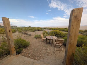 Una terraza de madera con una mesa y sillas, rodeada de vegetación desértica y un cielo despejado.