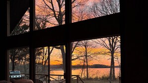 Un espace repas doté d’une grande table et de chaises, donnant sur un lac bordé d’arbres, avec une vue imprenable sur le coucher de soleil à travers de grandes fenêtres.