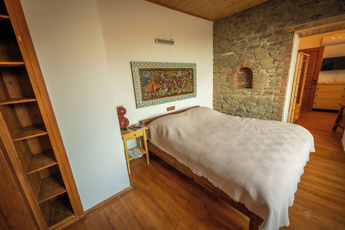A bedroom with a wooden bed, a nightstand, a stone wall, and a wooden staircase.
