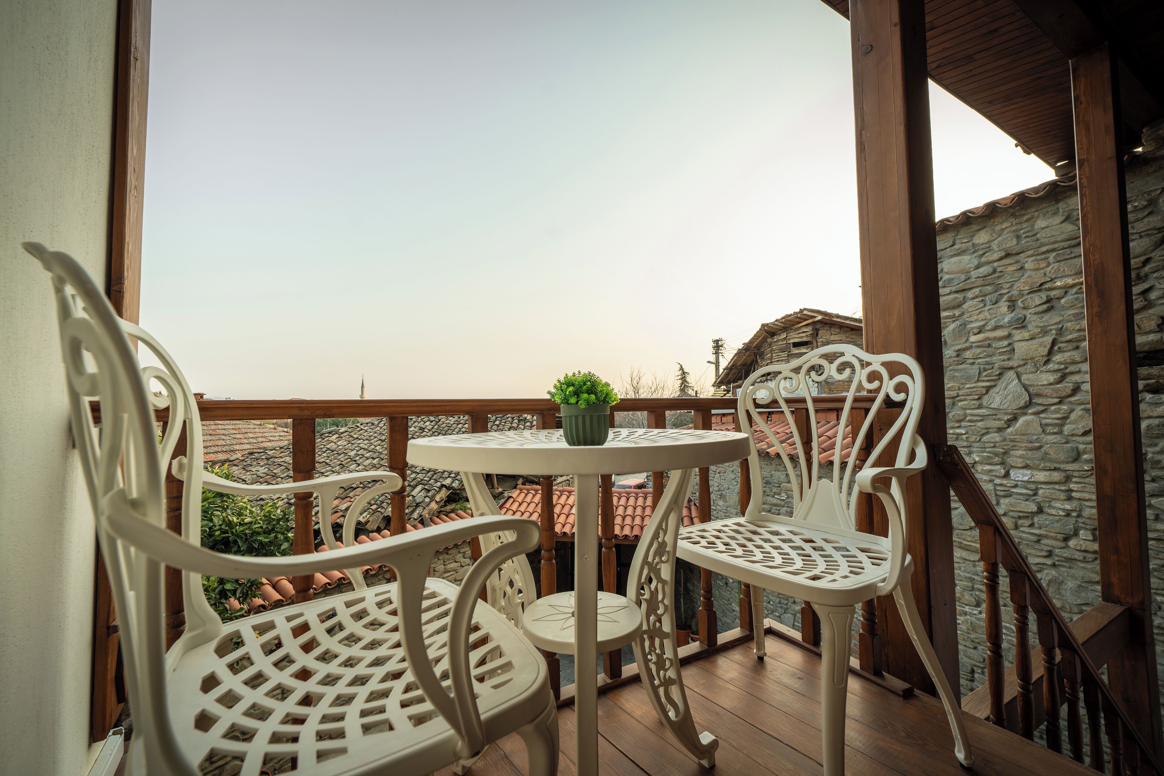A balcony with two white metal chairs and a small table, overlooking a stone building and a tiled roof.