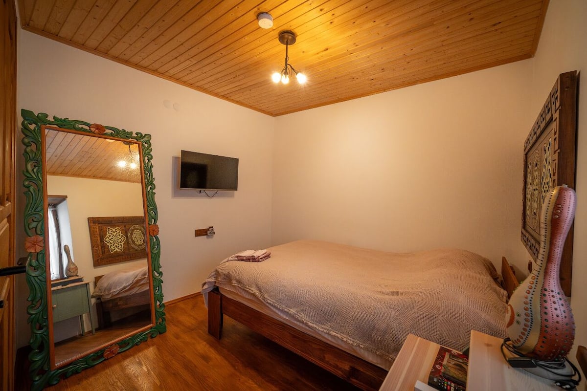 A bedroom with a wooden ceiling, a bed, a mirror, a television, and a decorative wall piece.