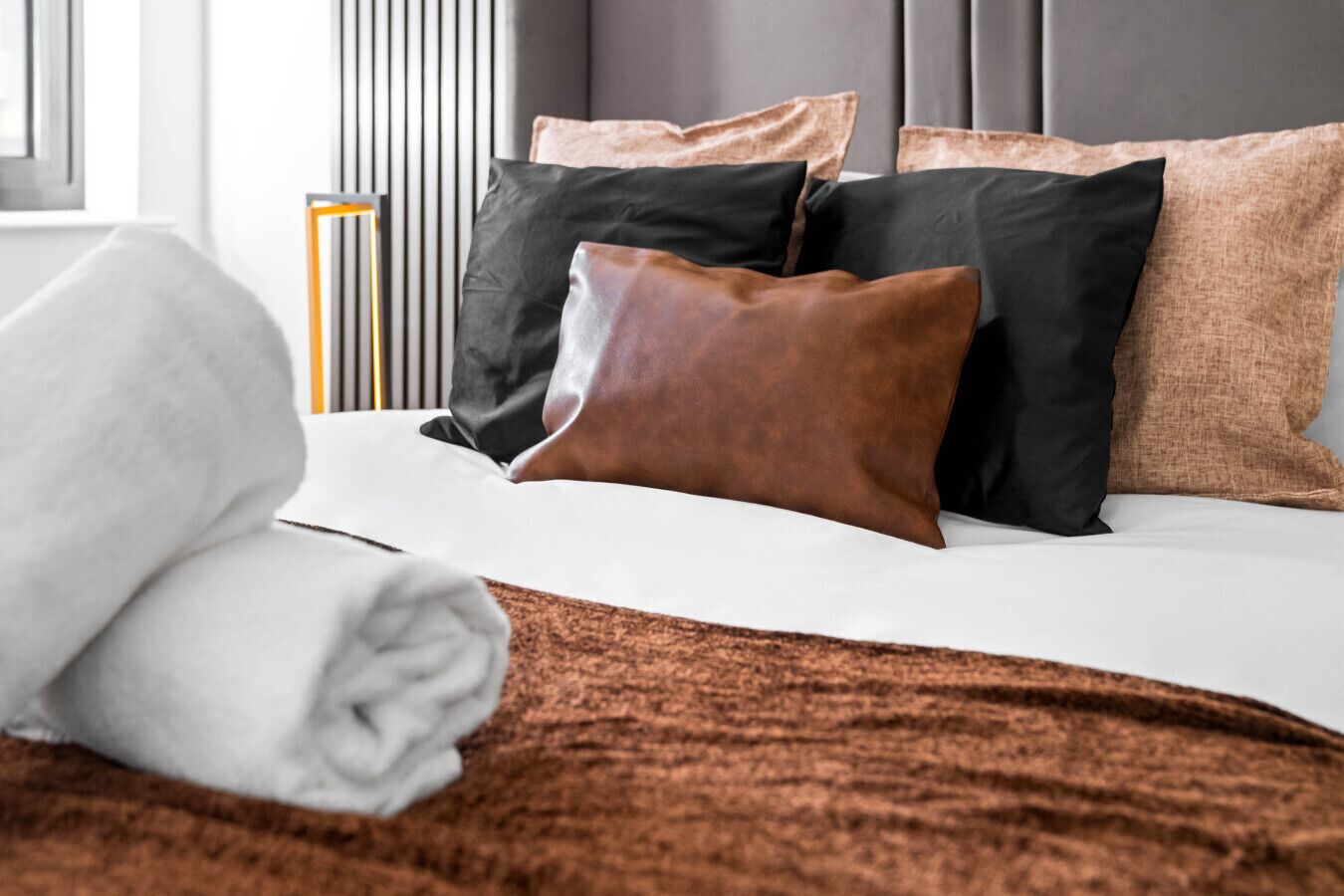 Un lit bien fait, avec une couverture marron, des oreillers noirs et marron, et une serviette blanche.