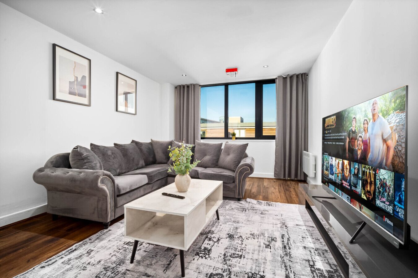 A modern living room with a grey sofa, a white coffee table, and a flat-screen TV displaying movie posters.