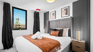 A modern hotel room with a large bed, a nightstand, a bedside lamp, a window with a view of a building, and a ceiling light. - Luxury 2 Bed Apartment (Birmingham)