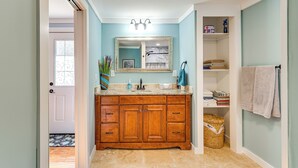 Un baño con tocador de madera, un lavabo y un espejo. Hay un perchero en la pared y una cesta en el suelo.
