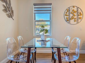 A dining area with a glass table, four clear chairs, a potted plant, and a window with blinds.