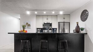 Une cuisine moderne avec un plan de travail noir, des armoires blanches et des appareils en acier inoxydable. On y trouve trois tabourets noirs près du plan de travail et un élément décoratif mural au-dessus du réfrigérateur.