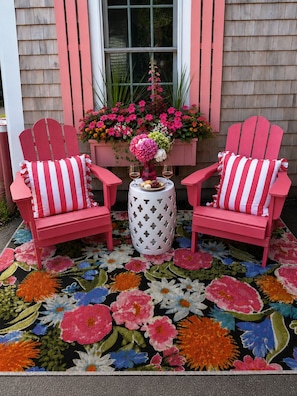 Two pink Adirondack chairs with striped cushions, a round table with a vase of flowers, and a floral-patterned rug.
