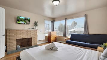 A hotel room with a brick fireplace, a bed with white bedding, a blue sofa, a television on the wall, and a window with curtains.