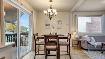 A dining area with a chandelier, a round table, and four chairs. There is a sofa and a window with blinds in the adjacent living area.