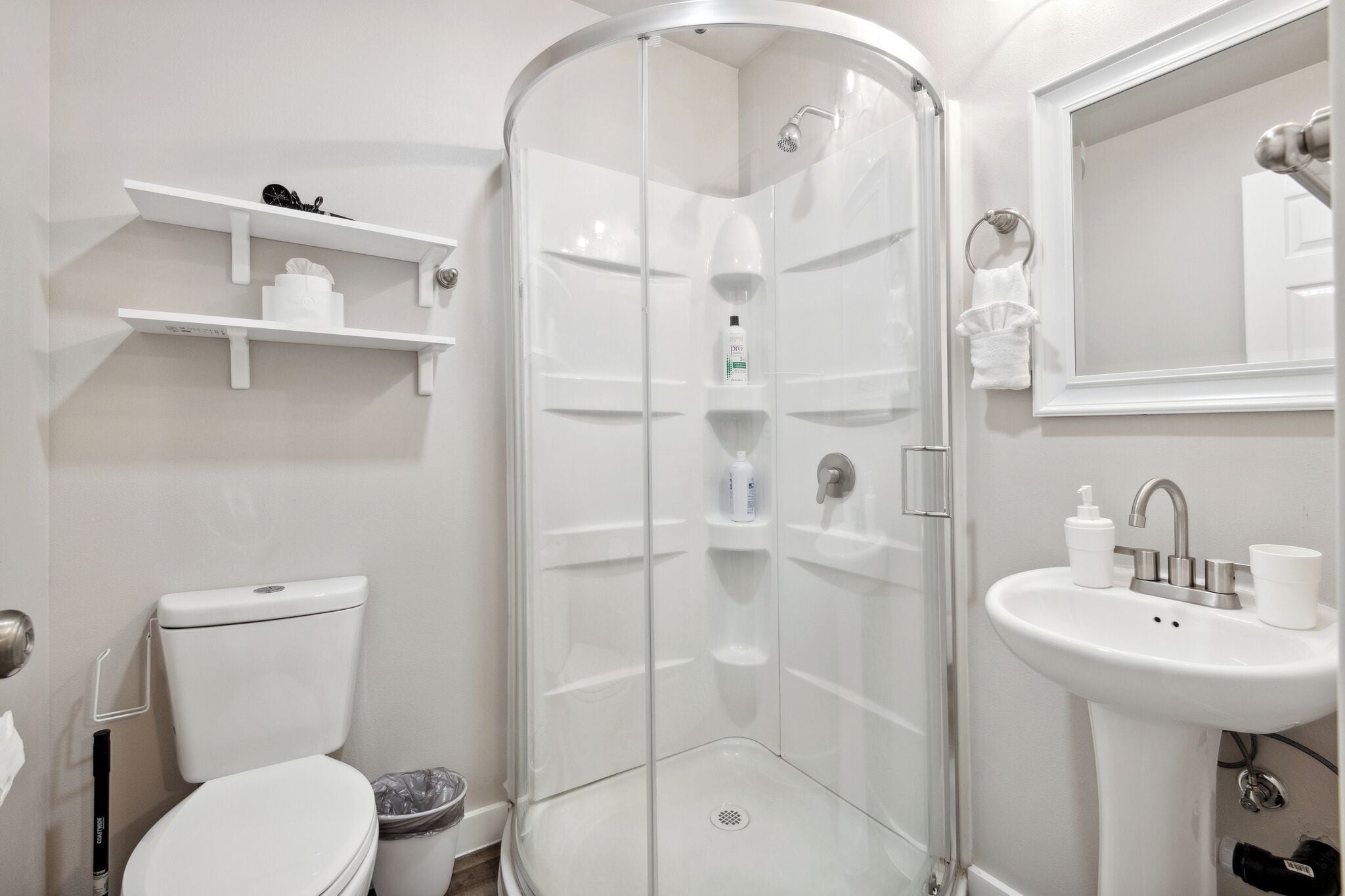 Une salle de bain moderne avec une douche à parois de verre, des toilettes, un lavabo avec un miroir et des étagères blanches où sont rangés les articles de toilette.