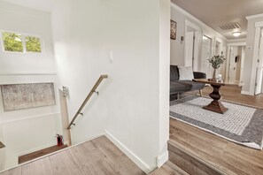 Un escalier avec une rampe en bois, un extincteur et un tapis gris menant à un espace de vie comprenant un canapé gris et une table basse en bois.