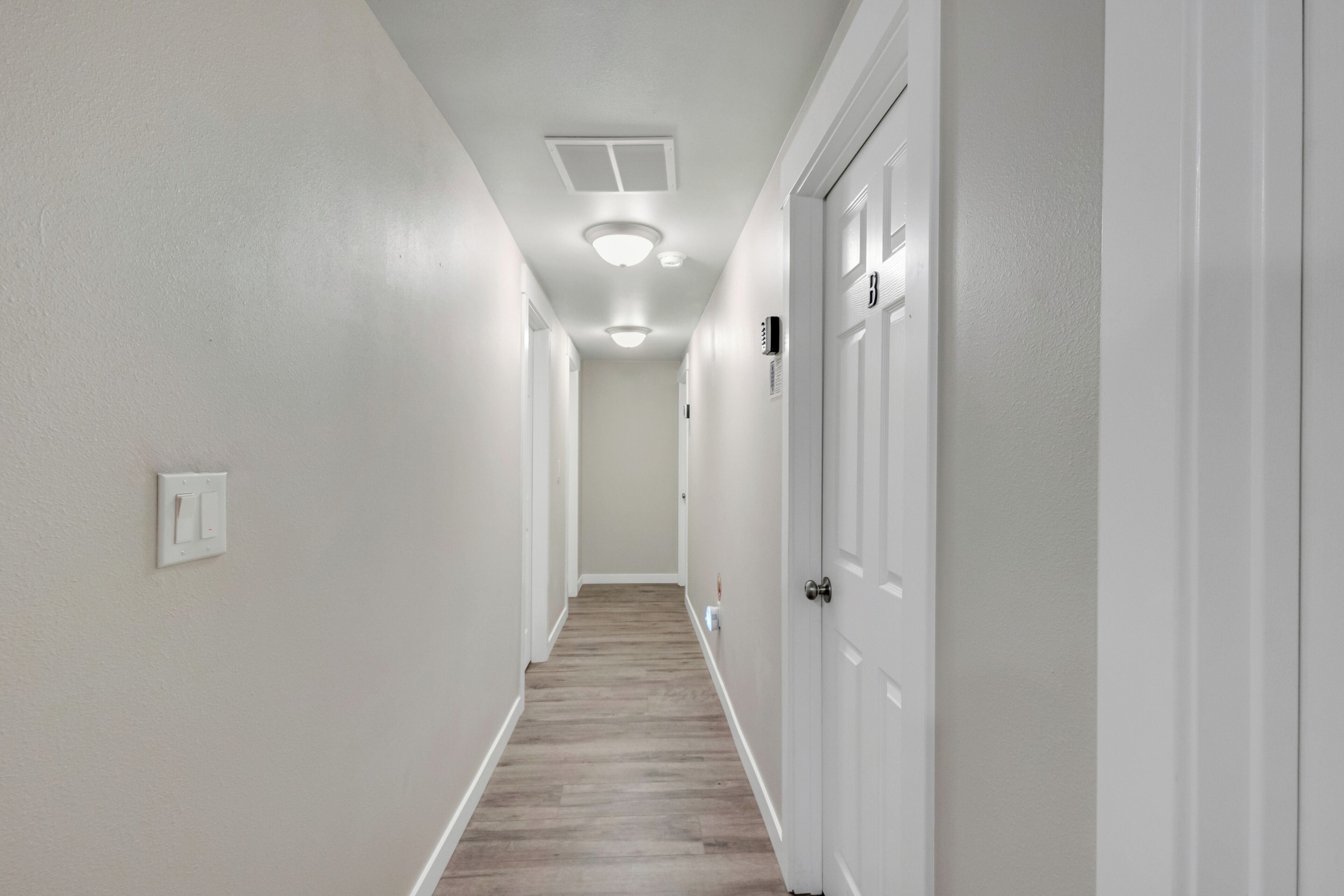 A narrow hallway with white walls and a wooden floor. There are white doors on the right and a light switch on the left.