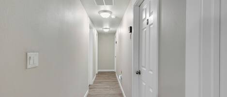 Un pasillo estrecho con paredes blancas y suelo de madera. Hay puertas blancas a la derecha y un interruptor de luz a la izquierda.