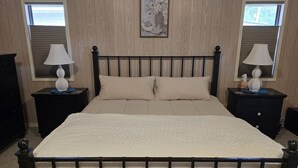 Una cama con cabecera, dos mesitas de noche con lámparas, un cuadro en la pared y ventanas con persianas.
