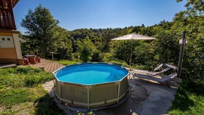 An outdoor swimming pool with a sunshade, surrounded by greenery and a patio area with lounge chairs.