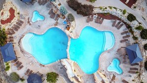 Vue aérienne d’un complexe hôtelier doté de deux grandes piscines, entouré d’espaces de détente agrémentés de parasols et de chaises longues.