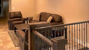 A living room with a brown leather sofa, two armchairs, and a coffee table. There are three windows on the wall, a staircase with a railing, and a patterned rug on the floor. - Peace at Zion + Zion Views + Resort Access (Orderville)